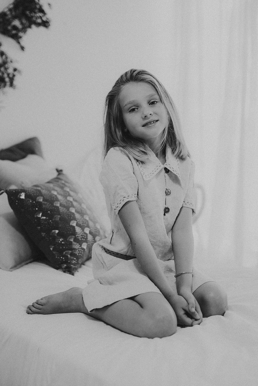 Black and white portrait of a young blonde girl sitting cross-legged on a bed, smiling at the camera.