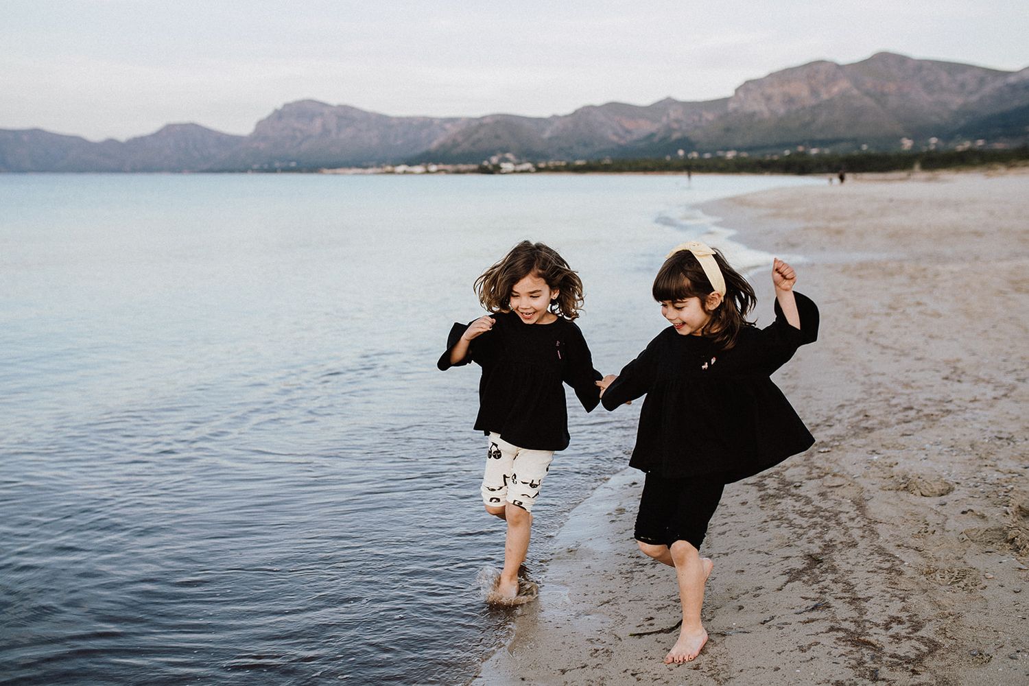 Two young girls holding hands and laughing while running along a sandy beach with mountains in the background.