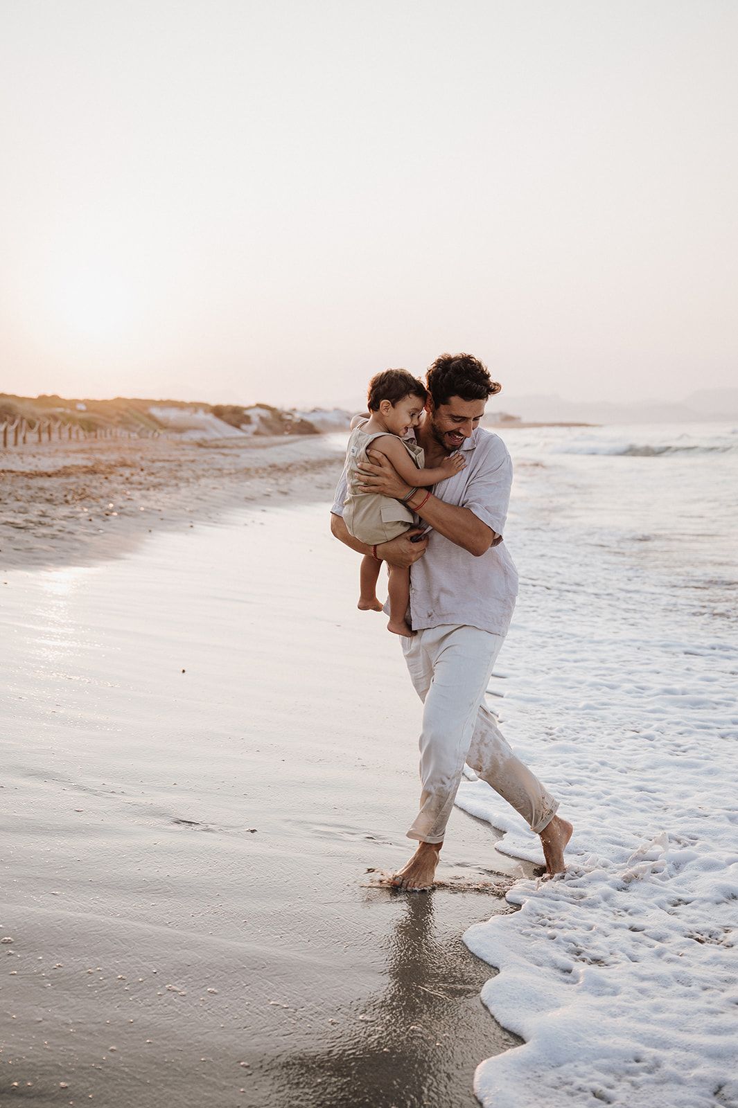 Father carries his toddler while walking barefoot along a scenic sunset beach.