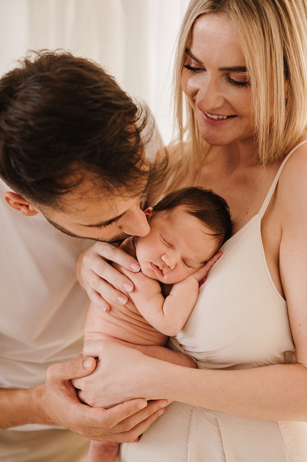 Happy parents kissing and holding their sleeping newborn baby close.