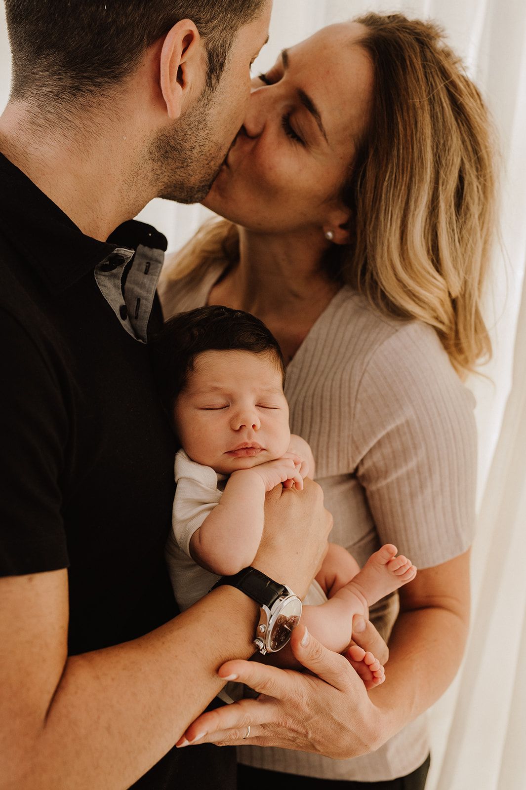 Parents sharing a kiss while cradling their sleeping newborn baby in a tender family portrait.