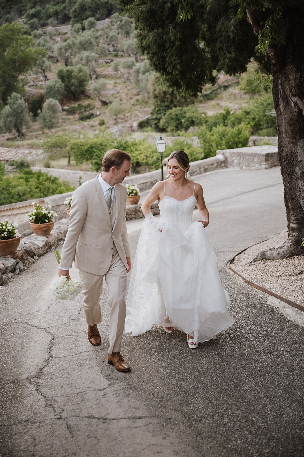 Bride in white ballgown and groom in beige suit walk together laughing on a scenic hillside road.