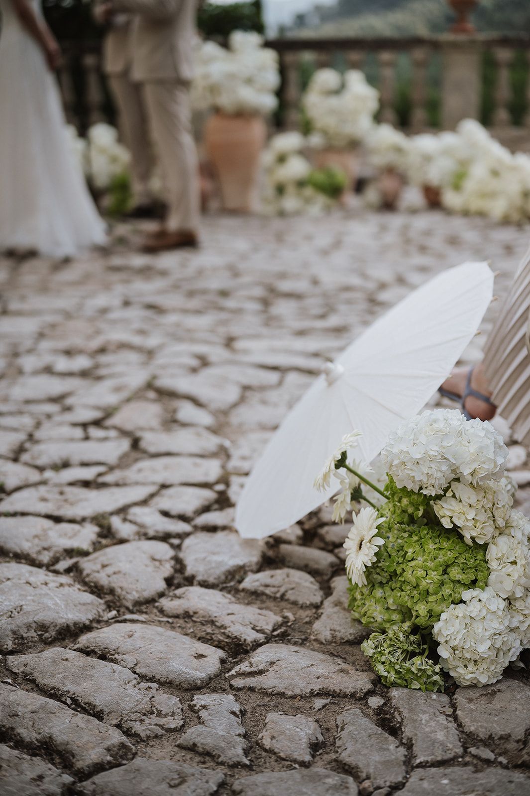 White paper parasol and green hydrangea bridal bouquet rest on cobblestone ground at outdoor wedding.