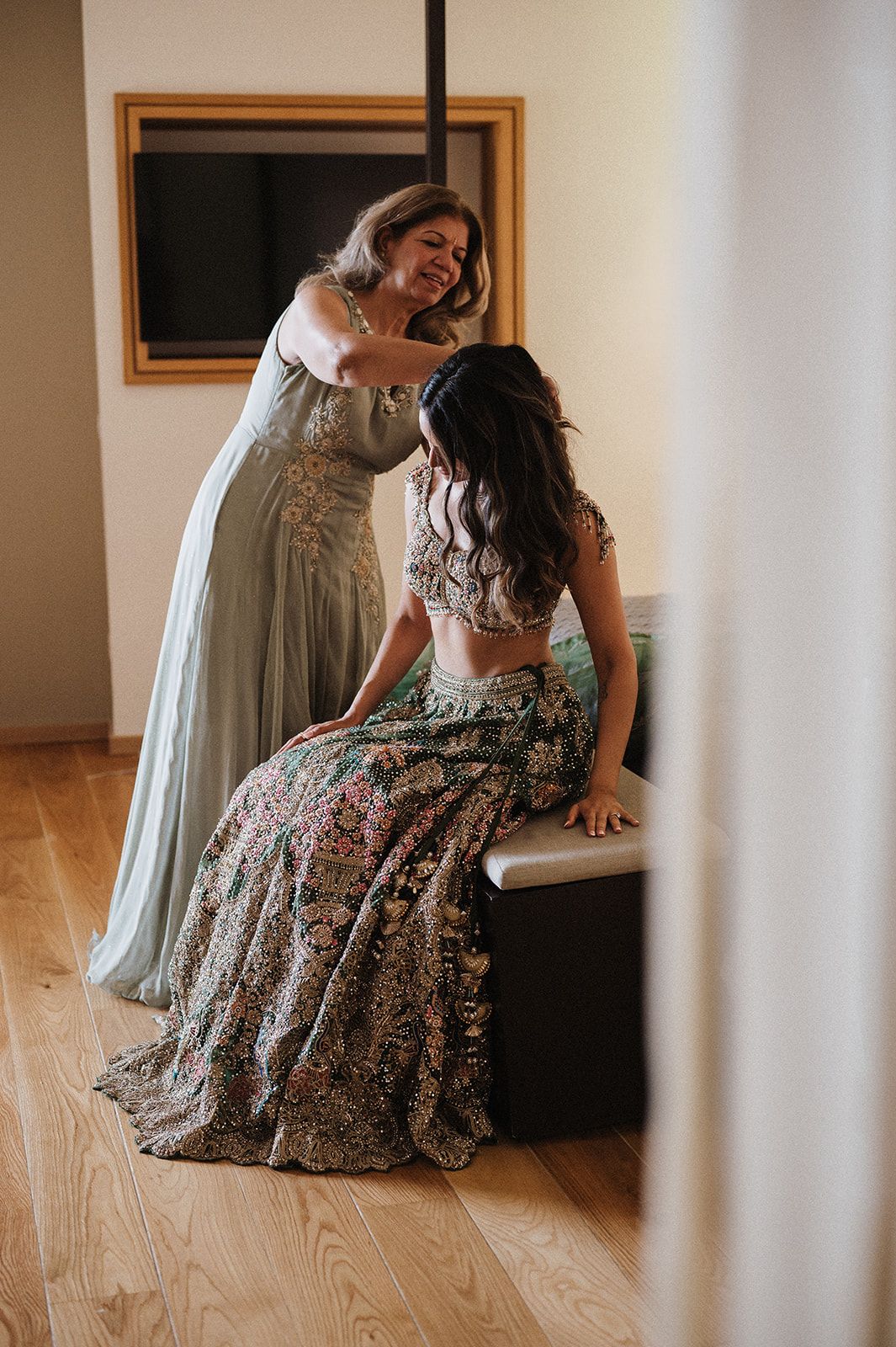 Mother helping bride in floral lehenga get ready in elegant hotel room before wedding.