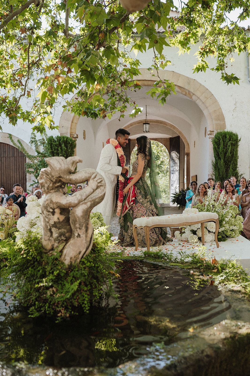 Couple sharing a kiss at outdoor Indian wedding ceremony with arched doorway and lush greenery.