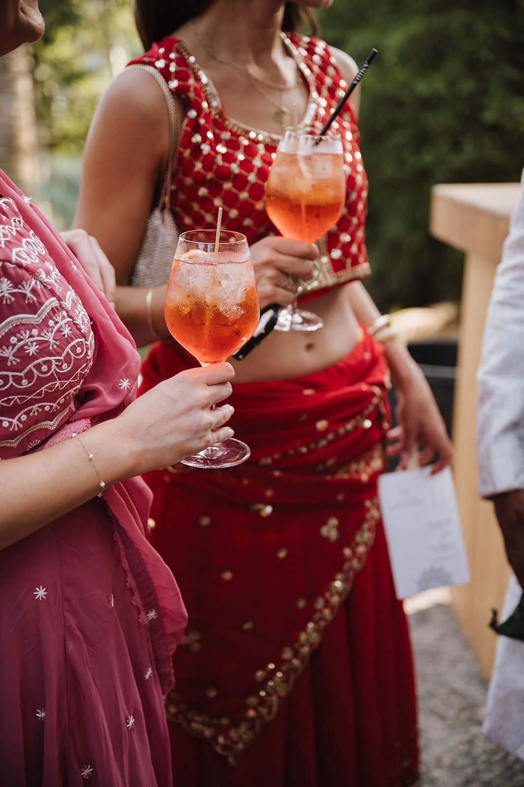Women in red and pink sarees holding orange Aperol Spritz cocktails at Indian wedding celebration.