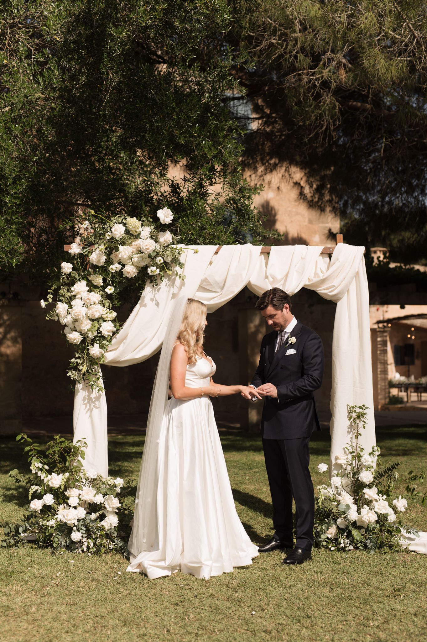 Bride and groom exchange rings under a floral arch draped with white fabric during an outdoor wedding ceremony.