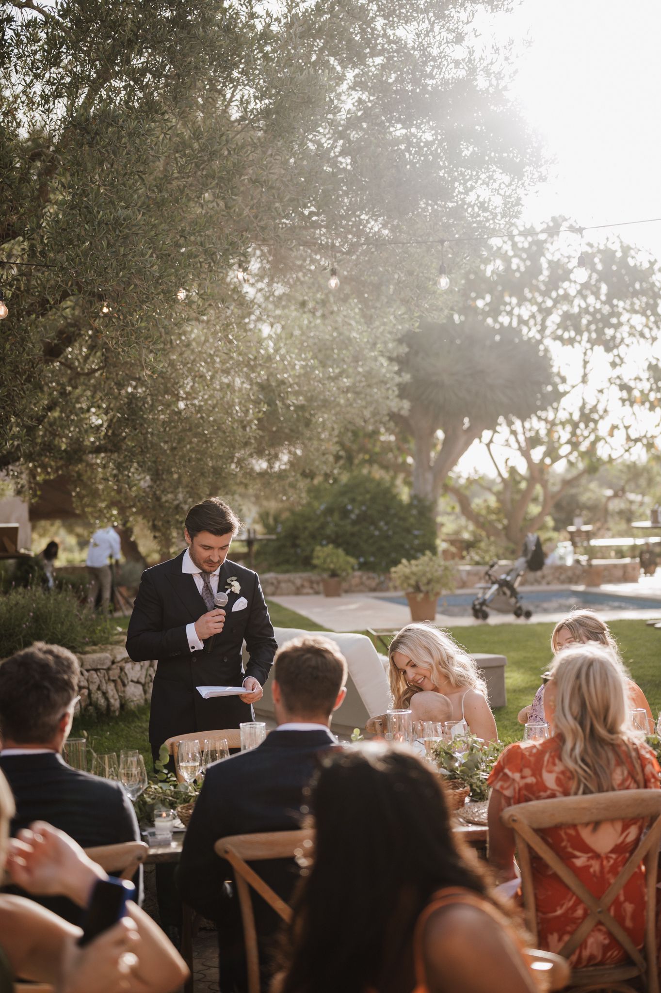 Groom delivering heartfelt speech at outdoor golden-hour wedding reception under olive trees.