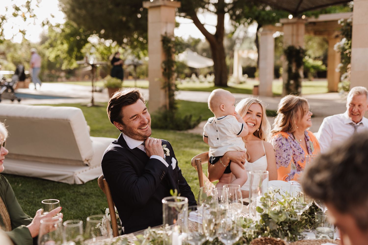 Bride and groom laughing joyfully with guests holding baby at outdoor wedding reception table.