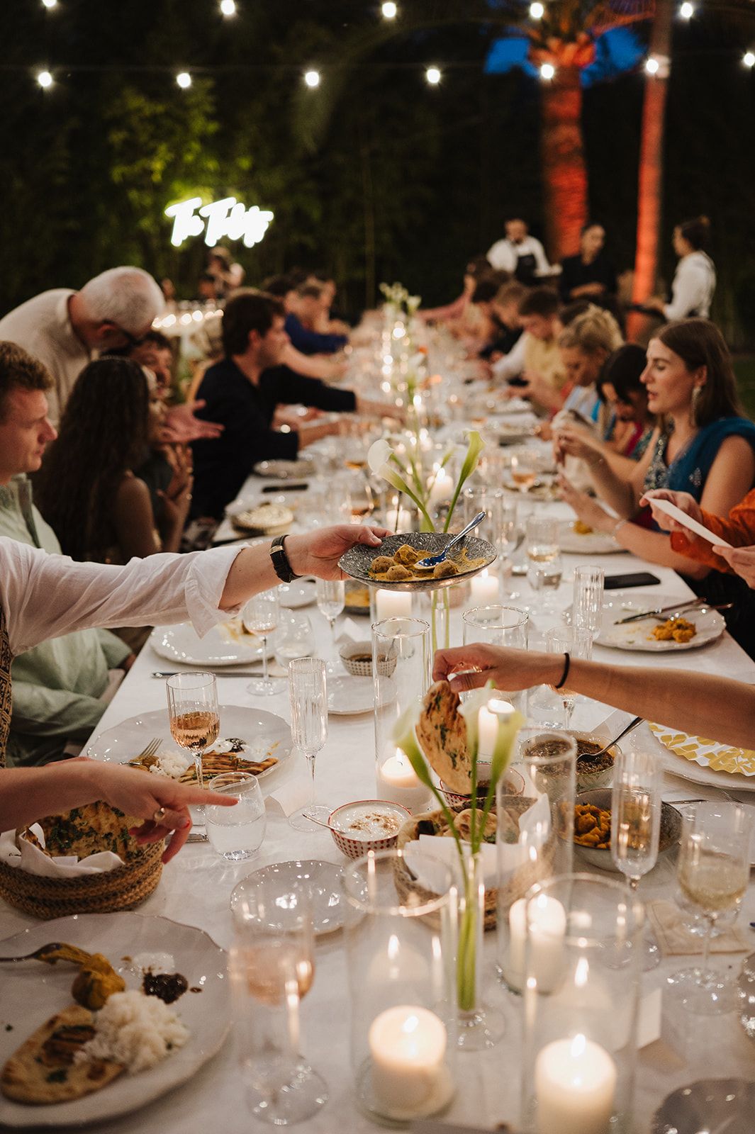 Guests share food along a long candlelit wedding reception table outdoors under warm string lights.