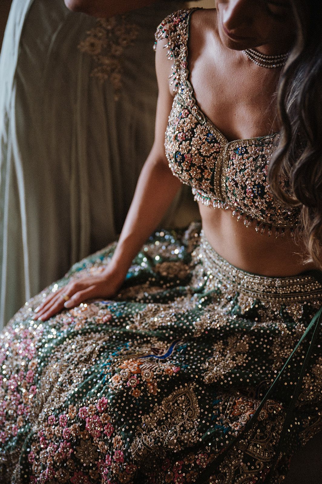 A bride wearing an ornate Indian lehenga with intricate beading, sequins, and floral embroidery in gold, pink, and teal tones.