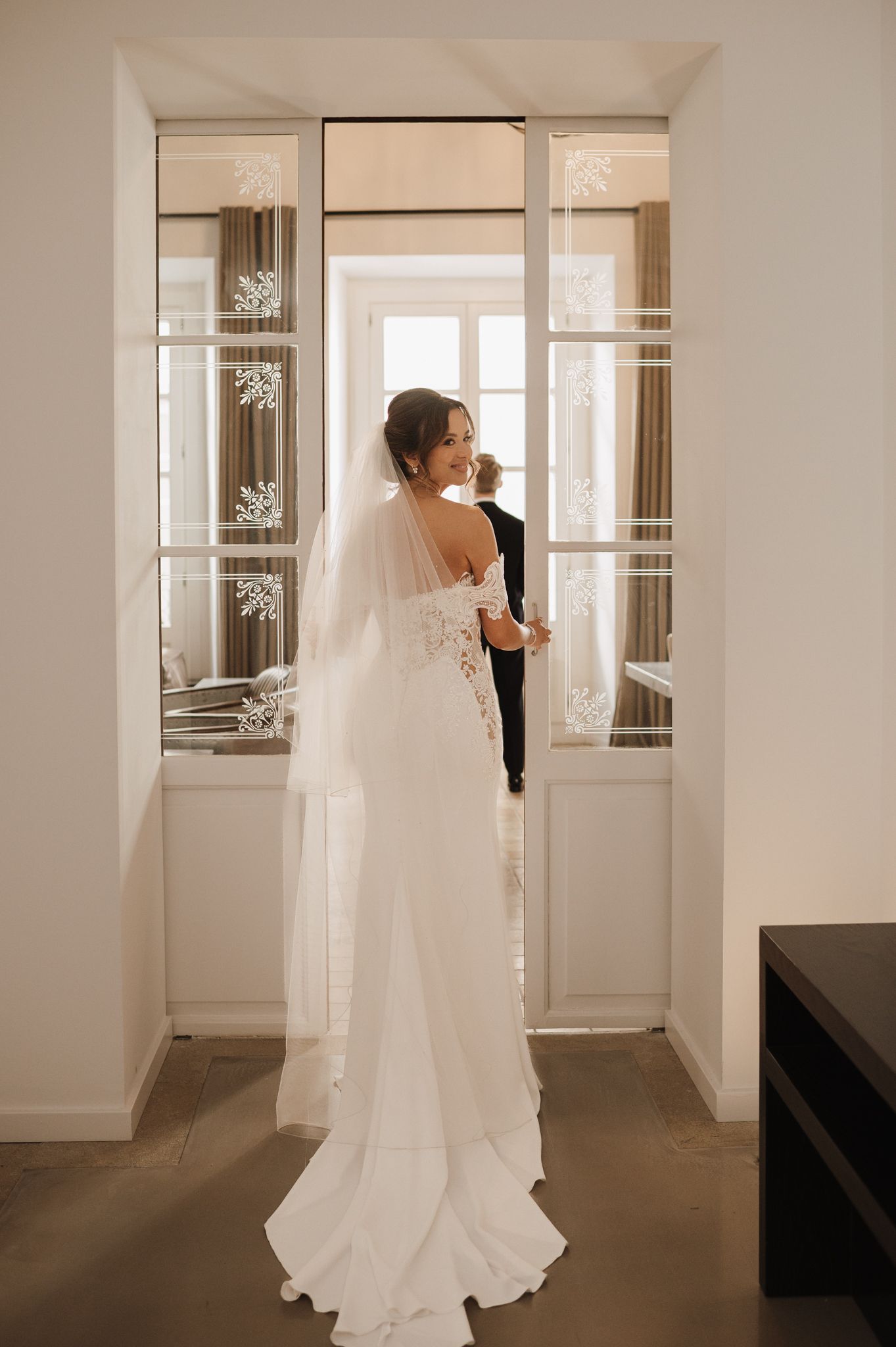 Bride in elegant lace backless wedding gown with veil standing by French doors in a bright white room.