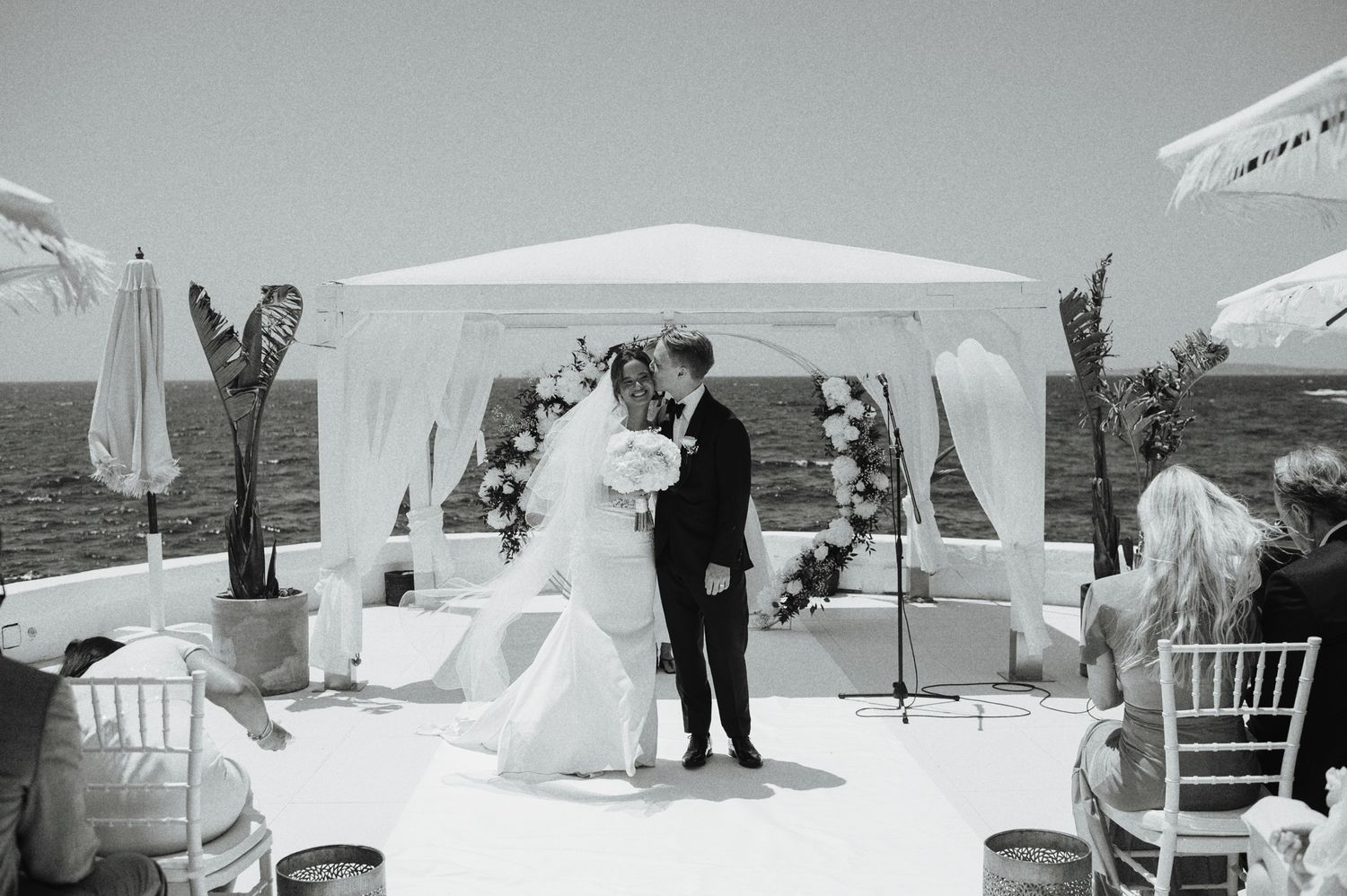 Black and white photo of bride and groom sharing first kiss at seaside wedding ceremony.