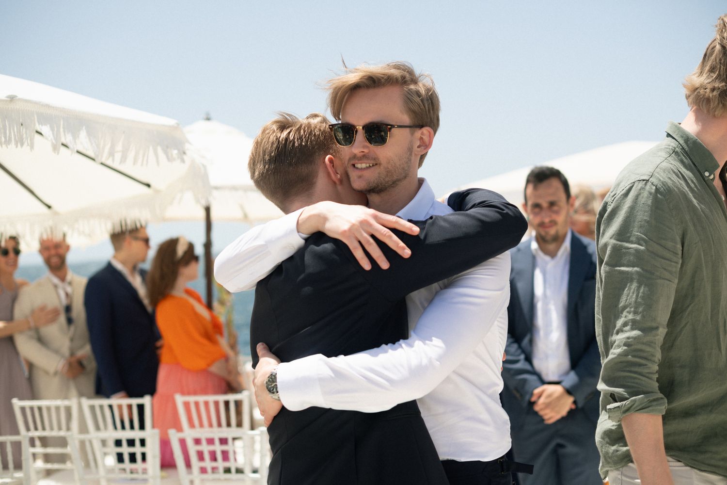 Two men share a warm embrace at an outdoor beachside wedding celebration on a sunny day.