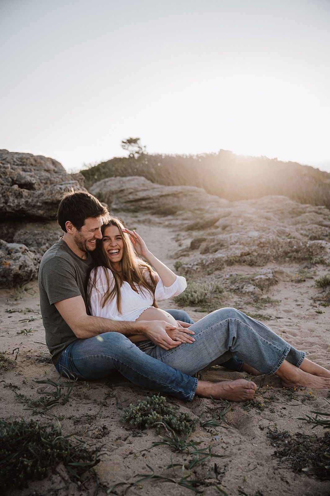 Couple sitting on sandy beach laughing, woman showing pregnant belly, golden sunset light over rocky landscape.