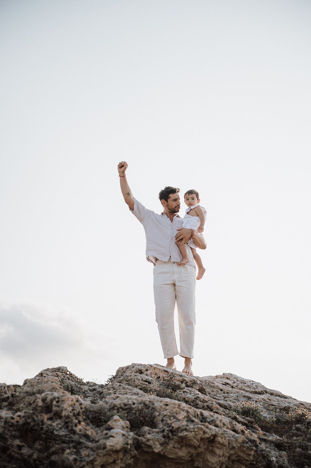 Father holding young child triumphantly on rocky outcrop, raising fist against a pale sky backdrop.