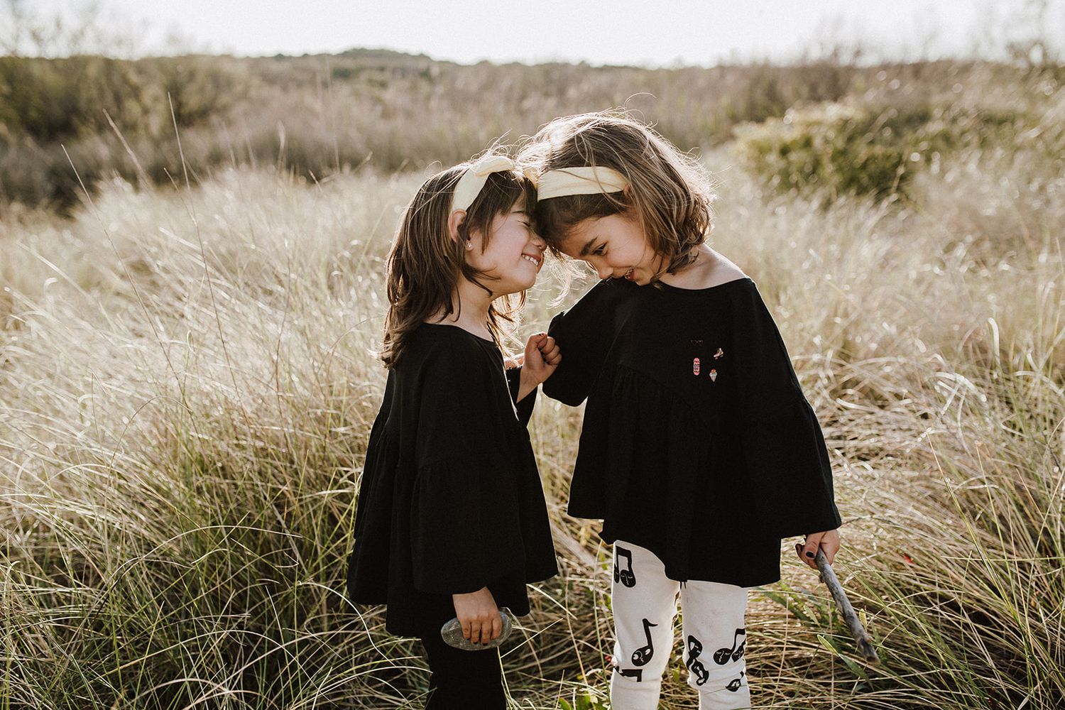 Two little girls in black outfits touching foreheads in a field of tall grass.