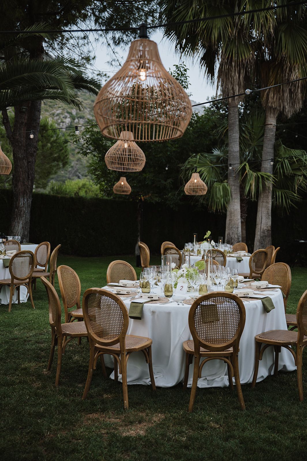 Elegant outdoor wedding reception with rattan pendant lights, round tables, white linens, and wooden cane chairs amid palm trees.