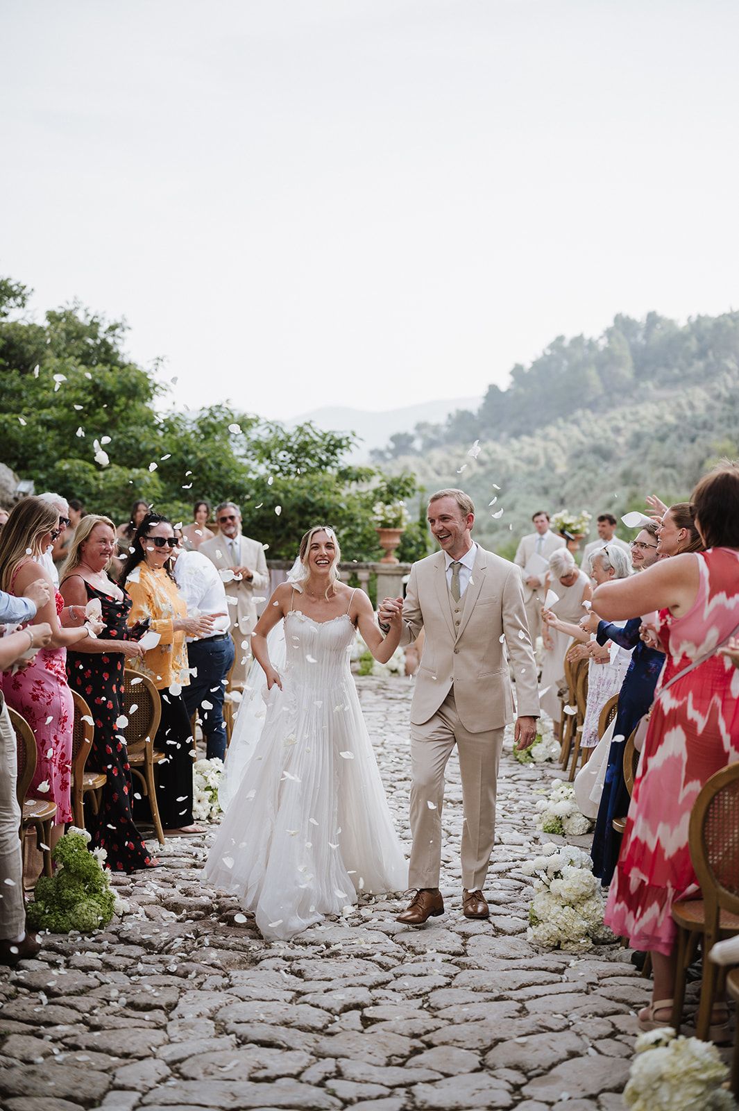 Newlyweds walk down cobblestone aisle as guests shower them with white flower petals after ceremony.