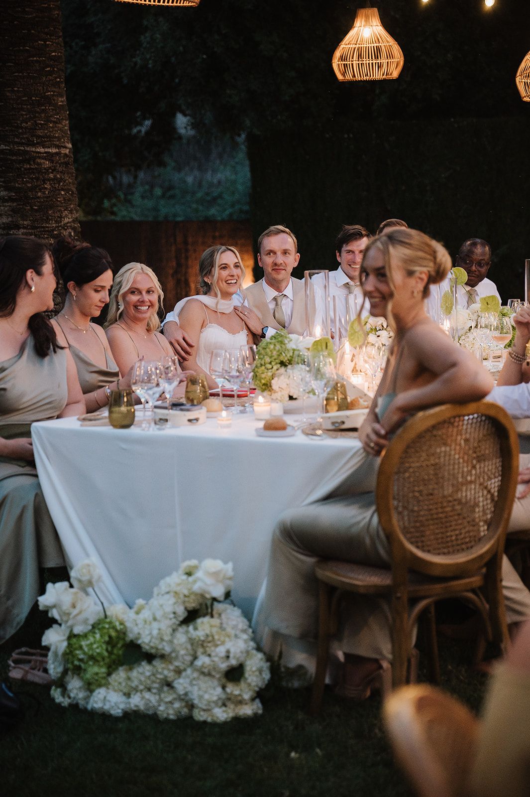 Bride and groom seated at elegant outdoor wedding reception table with bridesmaids and white florals.