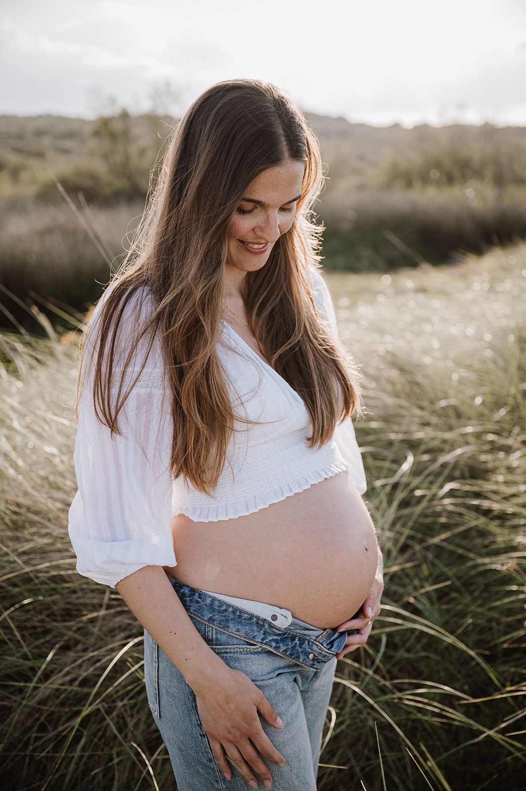 Pregnant woman in white crop top and jeans cradling baby bump in golden field during maternity photoshoot.