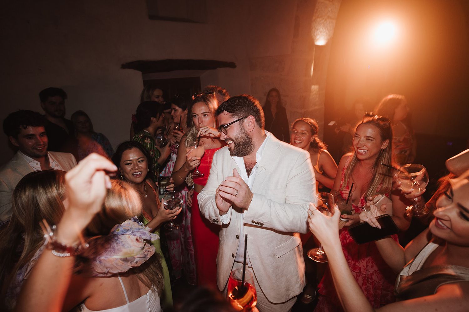 Groom in white suit celebrates joyfully with guests on the wedding reception dance floor.
