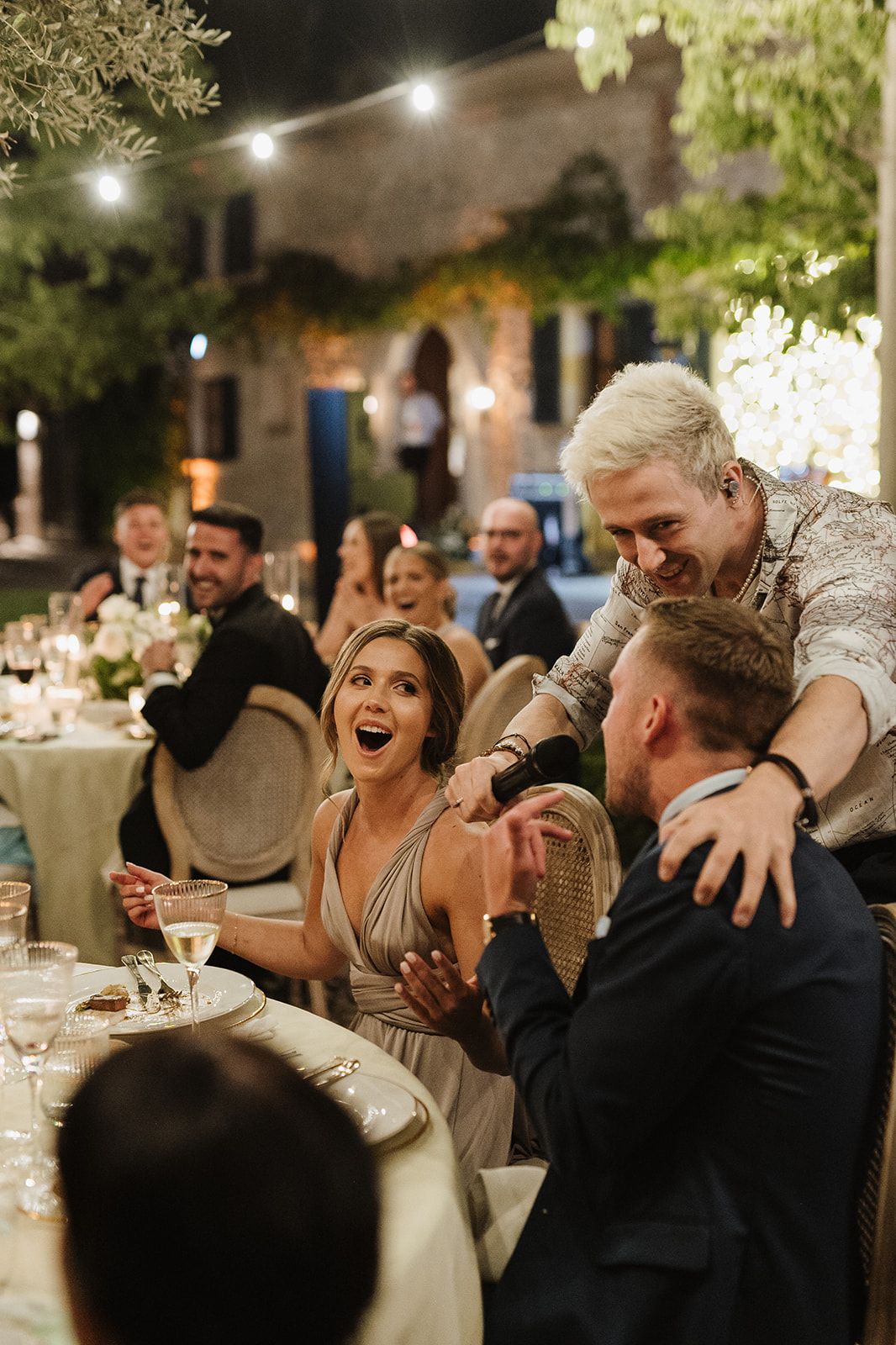 Wedding guests laugh and celebrate together at an elegant outdoor reception dinner under string lights.