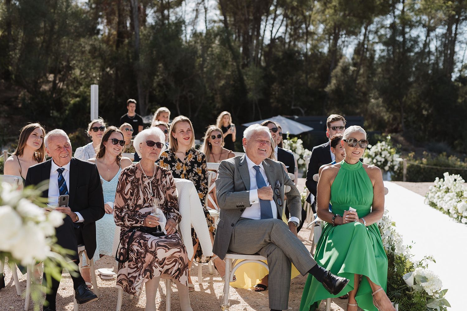 Wedding guests seated outdoors watching ceremony in sunny vineyard setting.