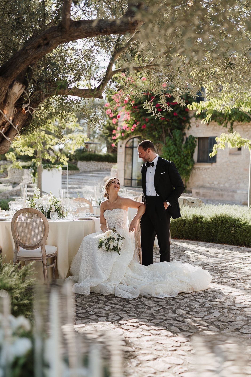 Bride and groom share romantic moment seated under olive tree at elegant outdoor wedding venue.