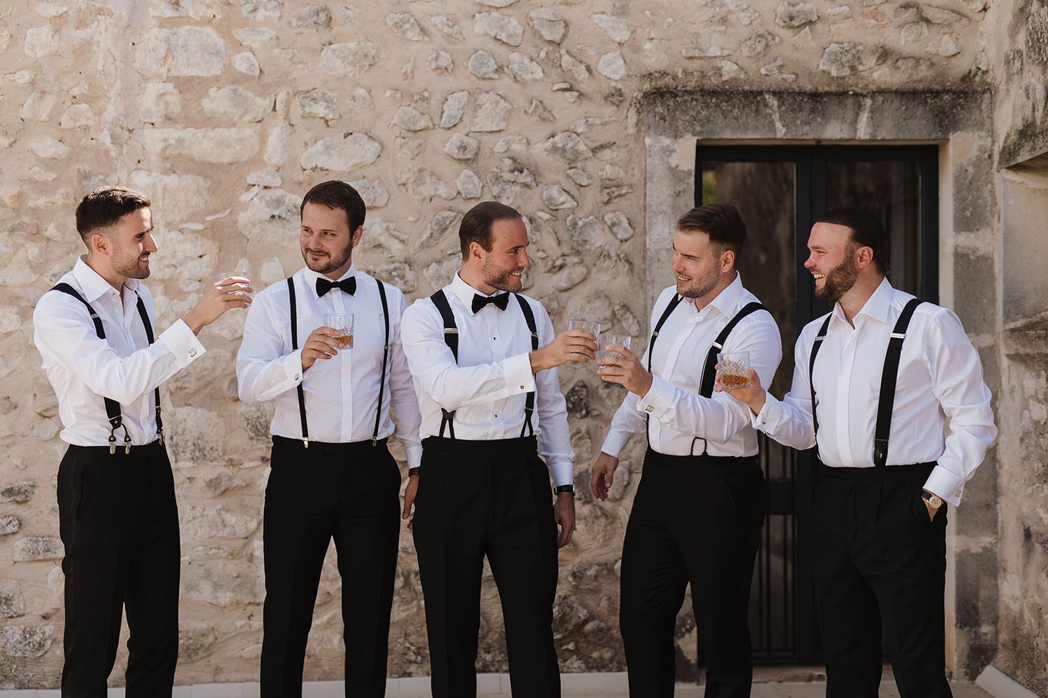 Five groomsmen in white shirts, black bow ties and suspenders laughing together against a stone wall.