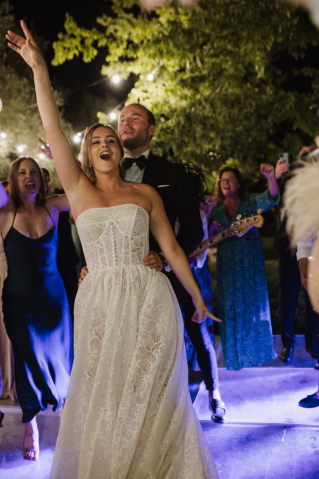 Bride in strapless lace gown raises her arm joyfully while dancing with groom at outdoor wedding reception.