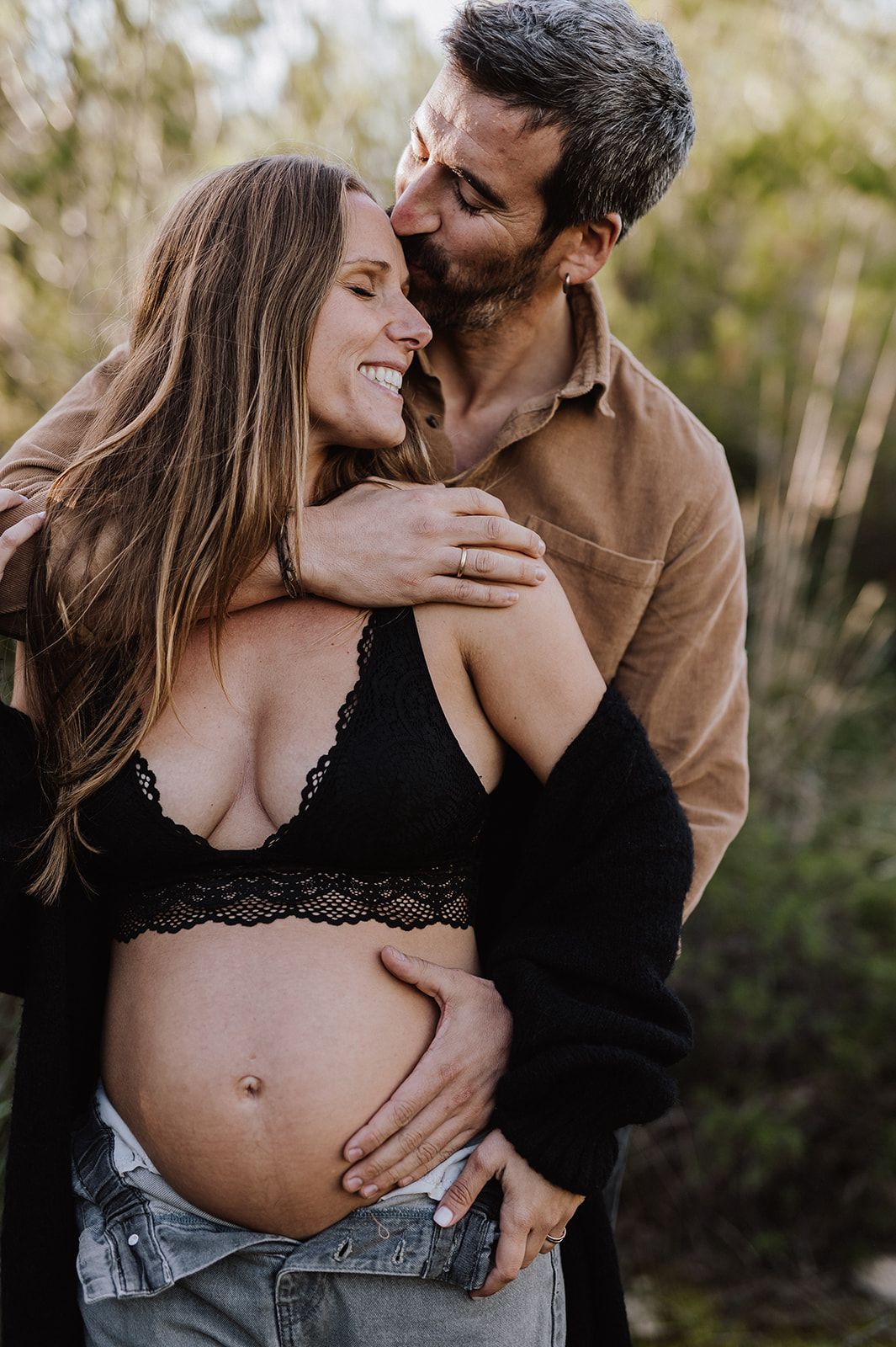 Smiling pregnant woman in black lace bralette embraced by her partner outdoors in a maternity photo shoot.