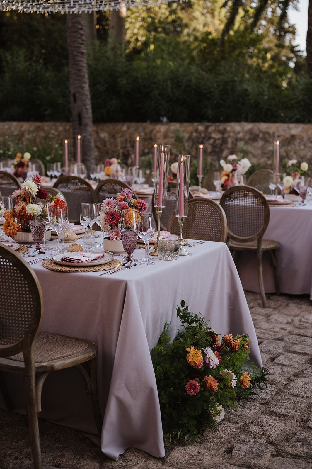 Elegant outdoor reception tables with pink linens, tall candles, colorful floral centerpieces, and rattan chairs on cobblestone.