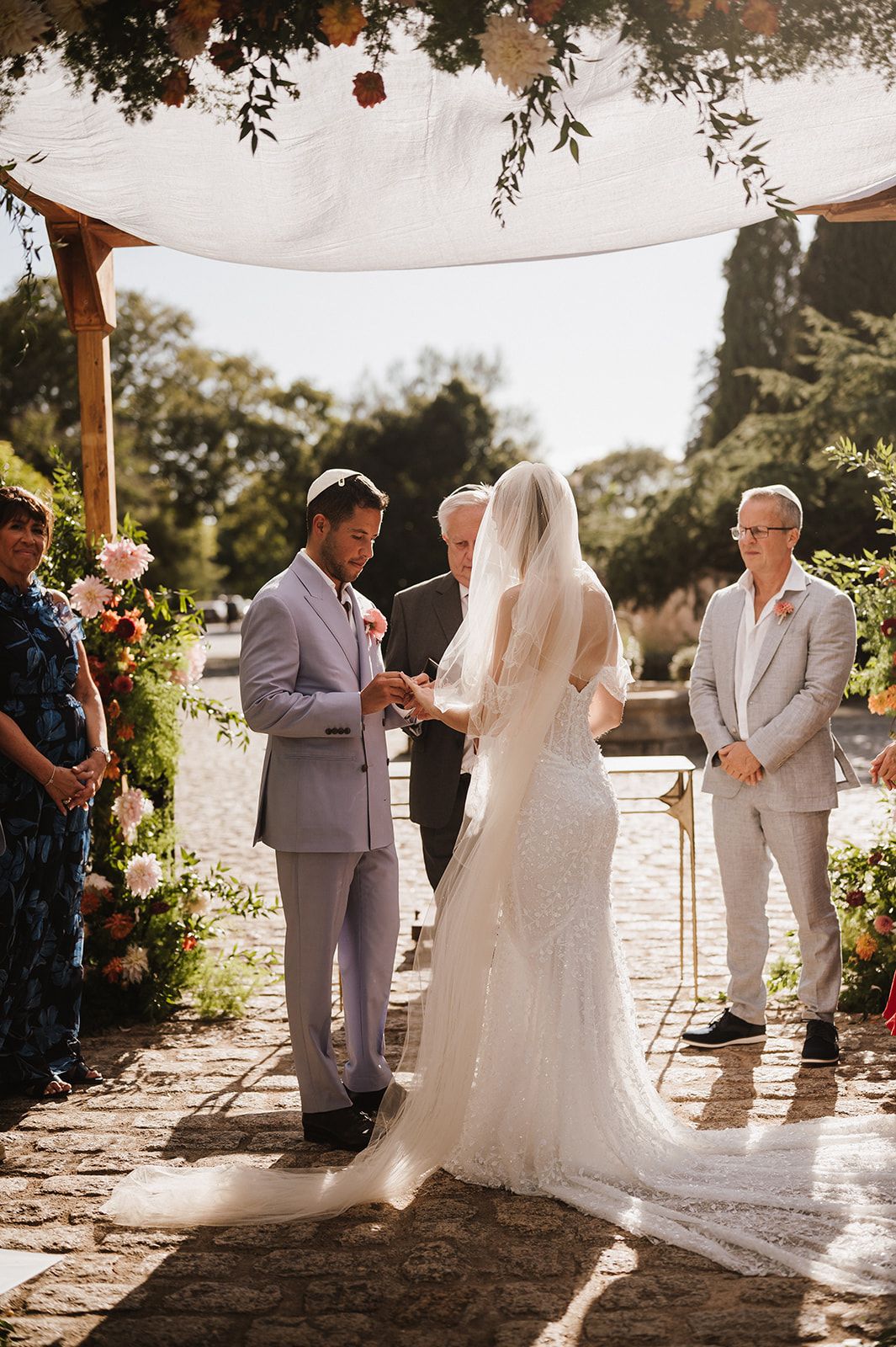 Groom places ring on bride's finger under floral chuppah outdoors.