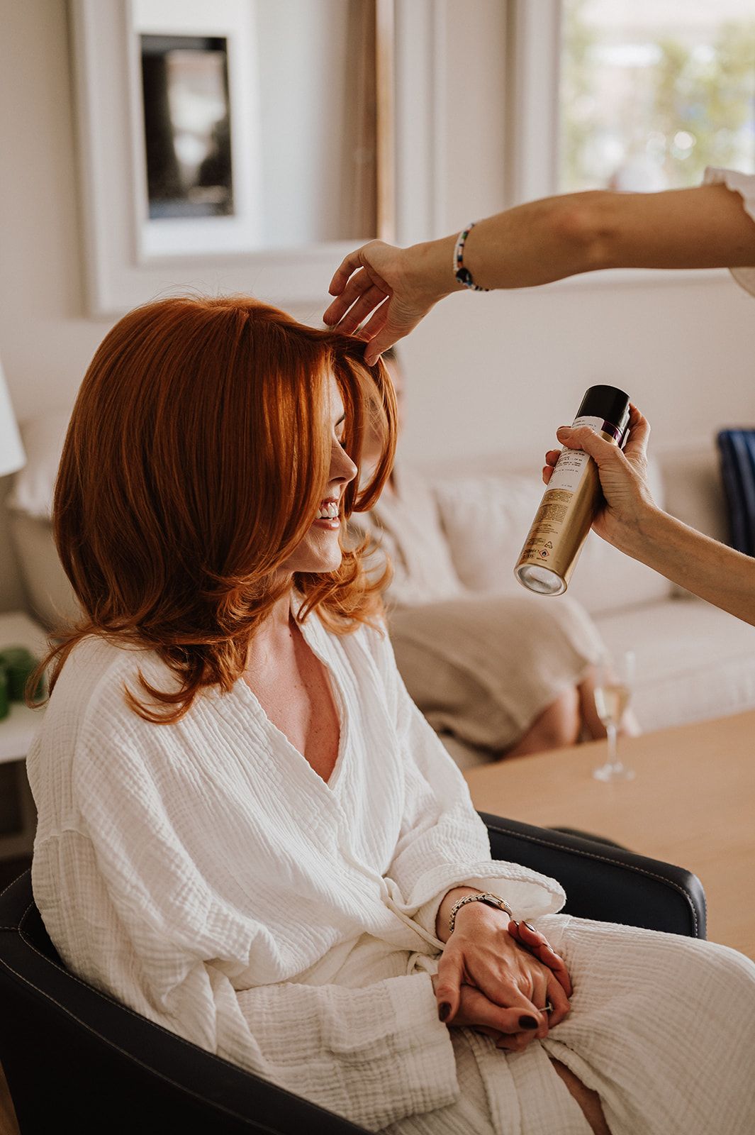 Bride with red hair getting hairspray applied by stylist while sitting in chair wearing white robe.