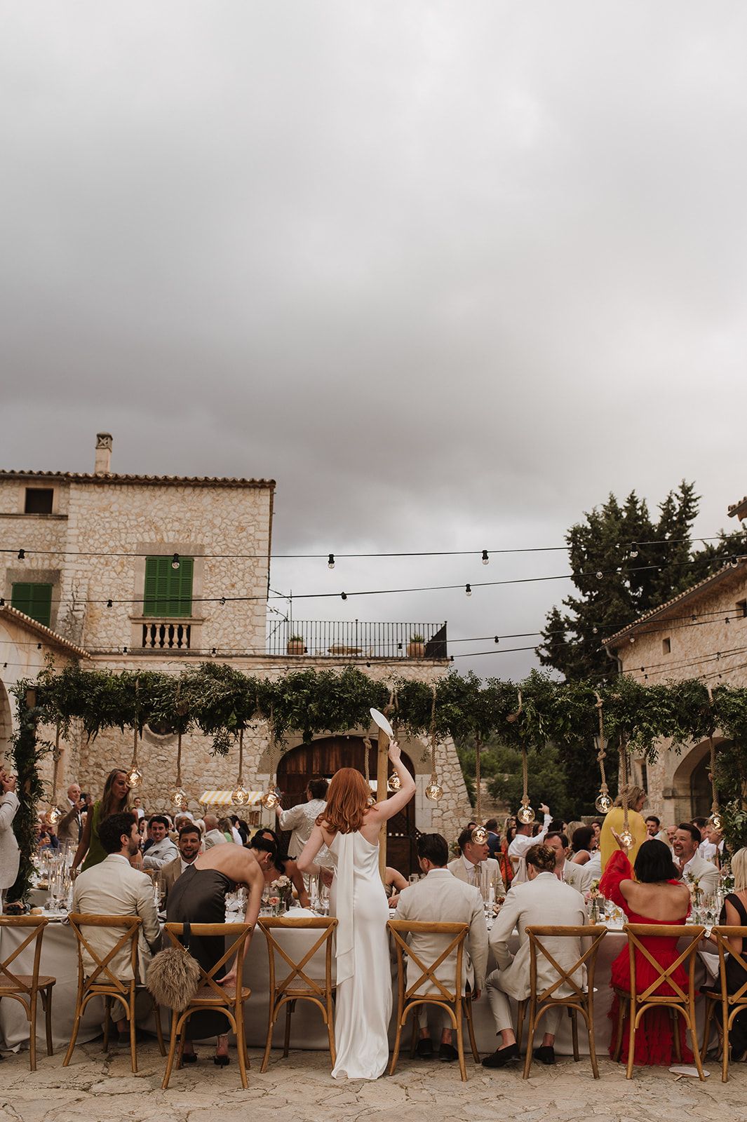 Elegant outdoor wedding reception at a stone villa with long dining tables, string lights, and overcast sky in Mallorca.