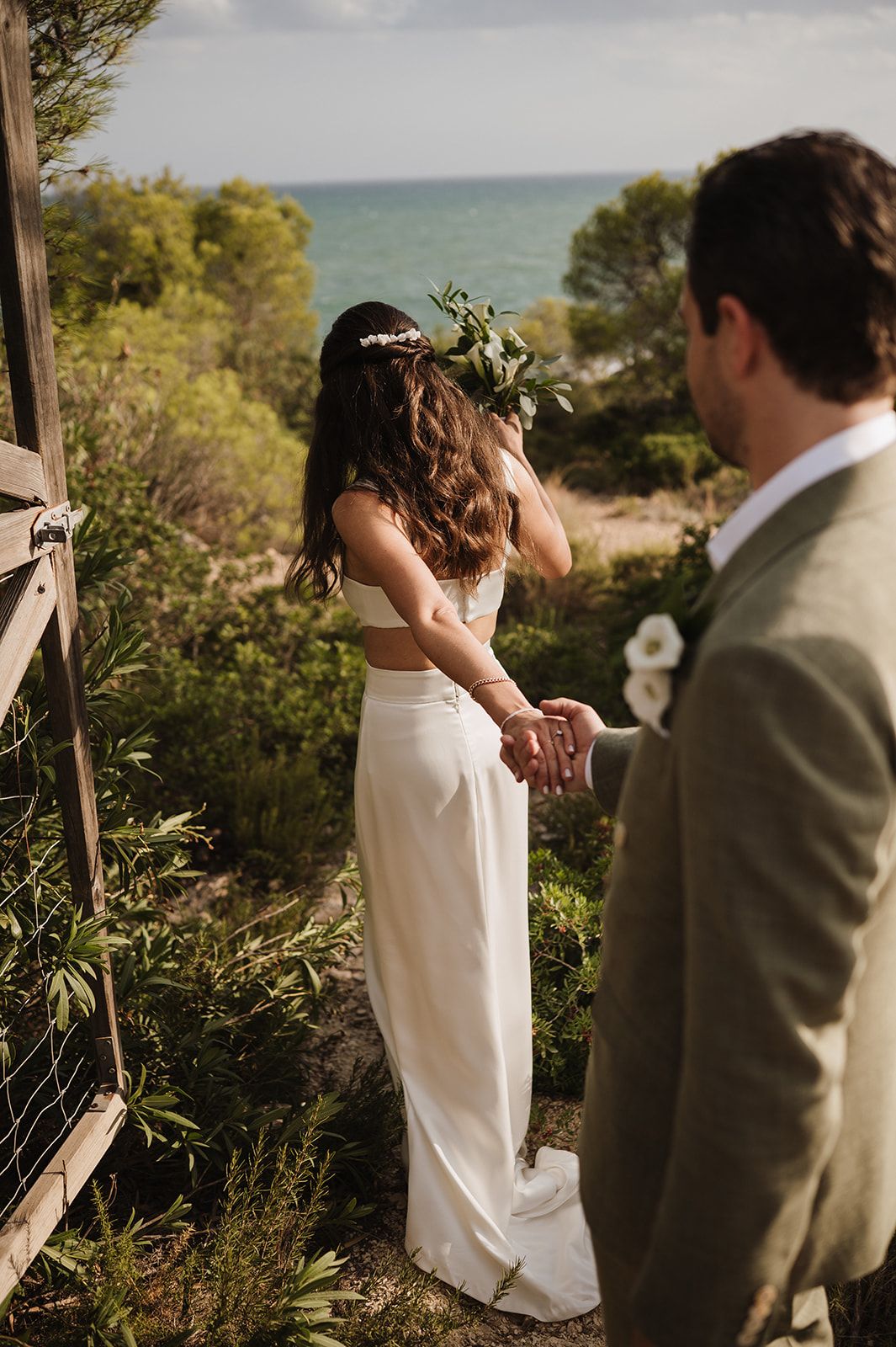 Bride in white gown with floral crown holds grooms hand overlooking ocean view.