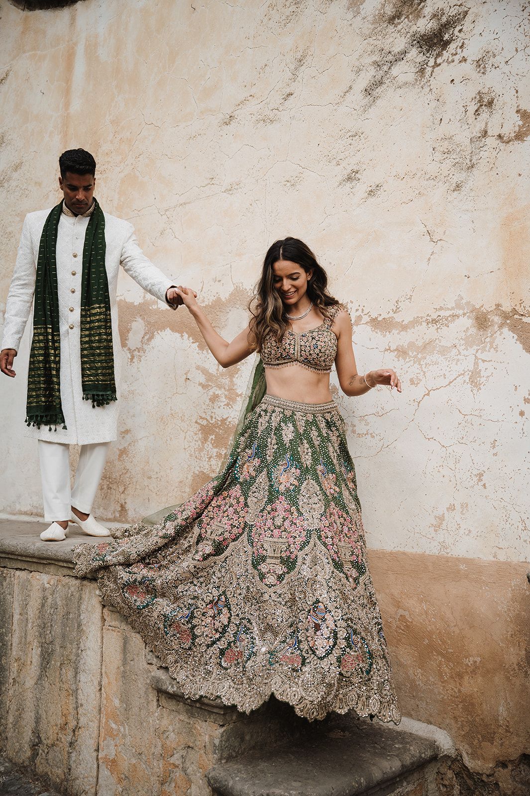 Couple in traditional Indian wedding attire posing against rustic wall outdoors.