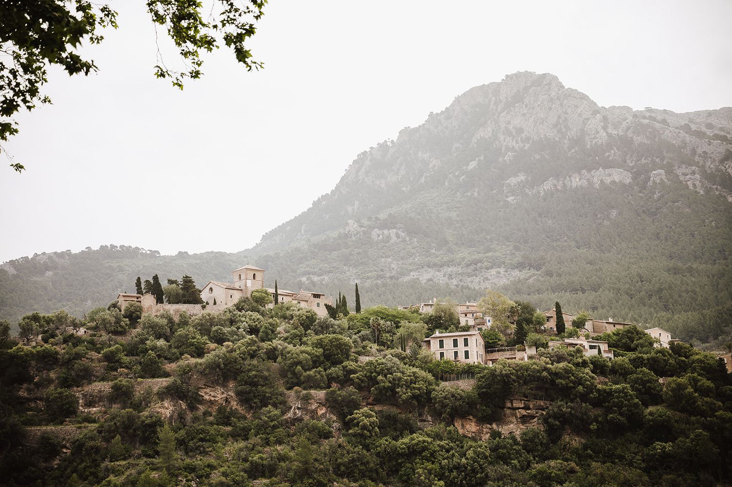 Scenic hilltop village nestled among lush green trees with dramatic rocky mountain backdrop in Mediterranean landscape.