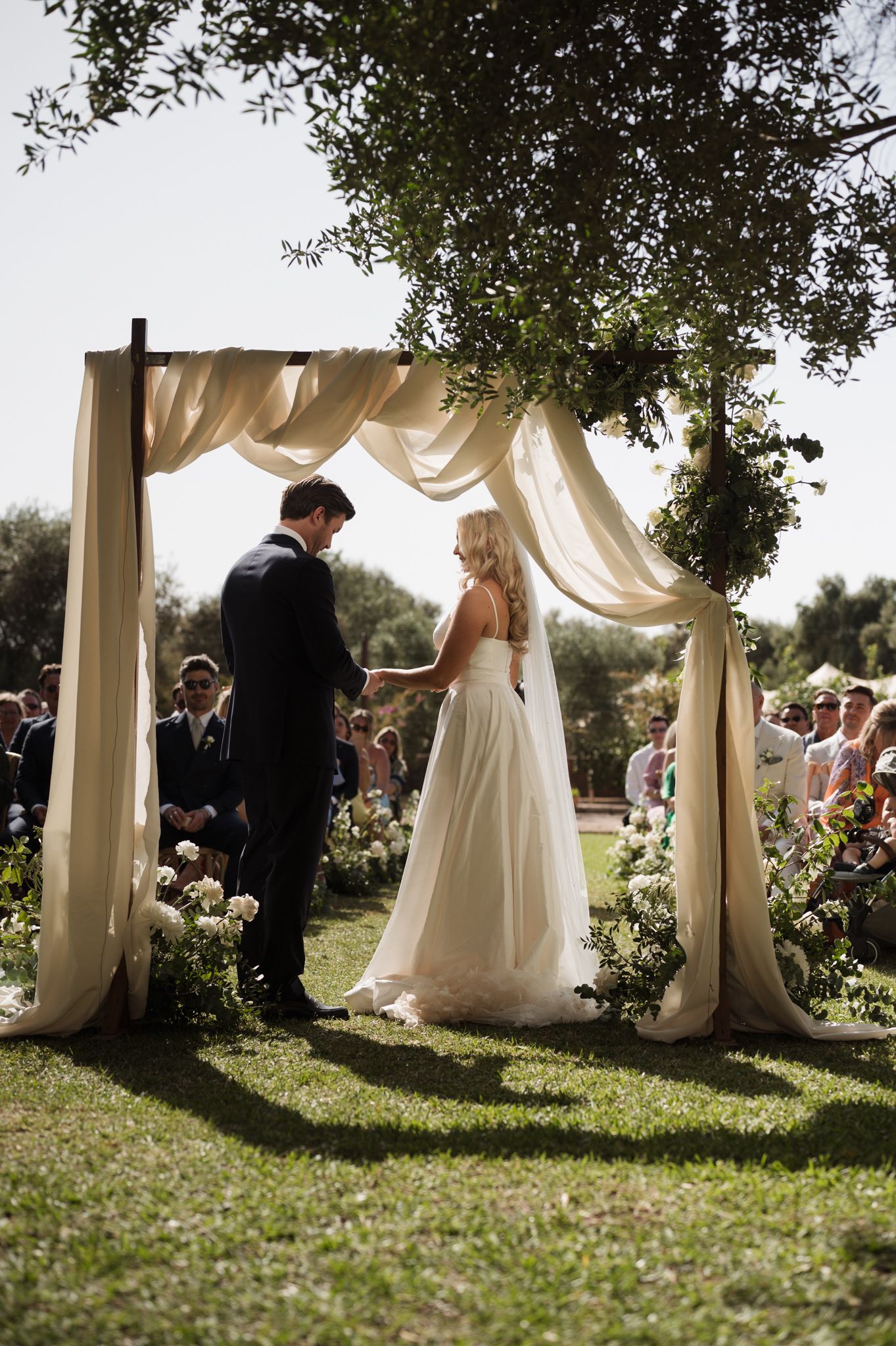 Bride and groom exchanging vows under draped arch with floral decor outdoors.