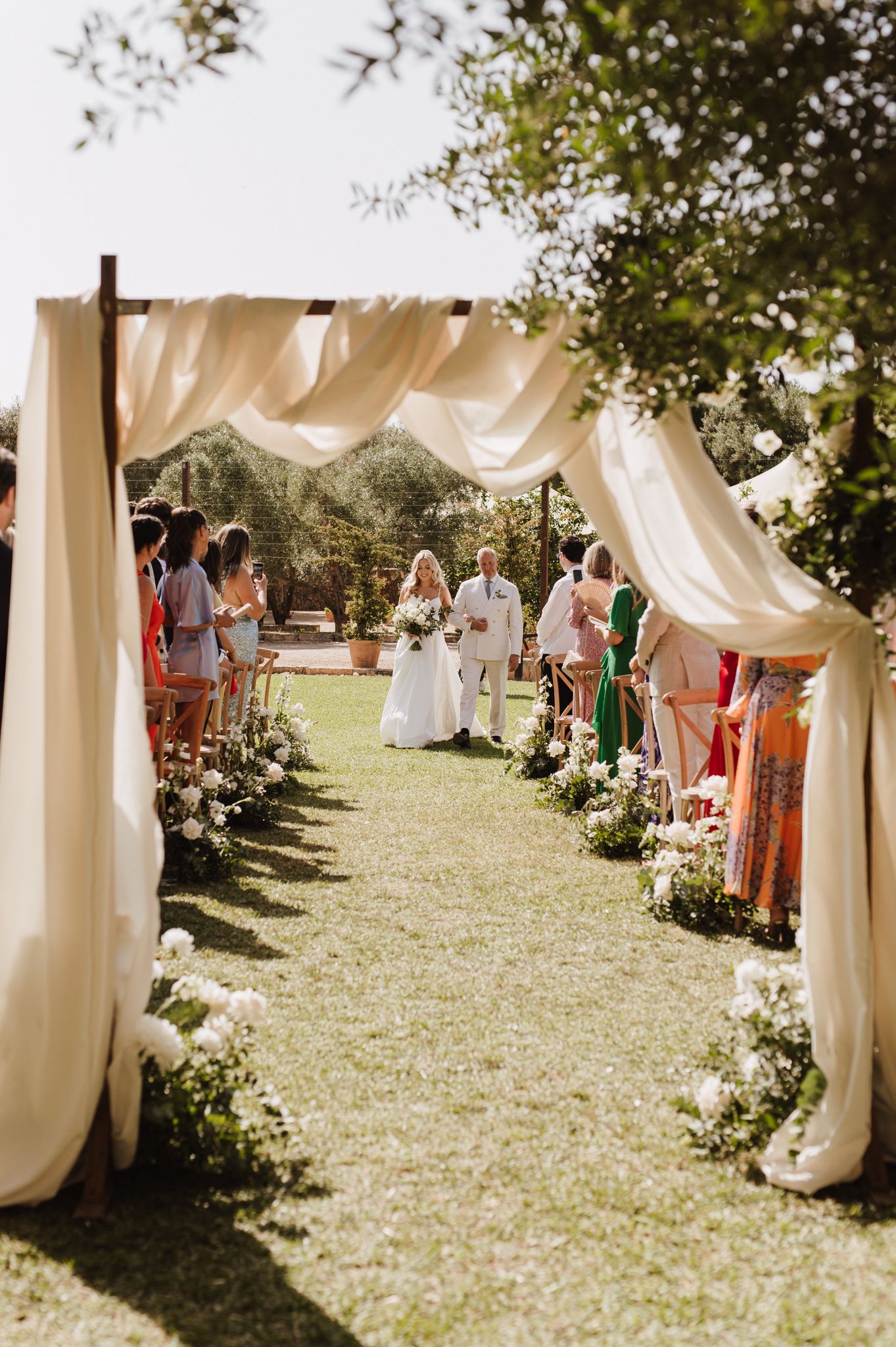 Elegant outdoor wedding ceremony aisle with white draping arch, floral arrangements, and guests lining the grass pathway.