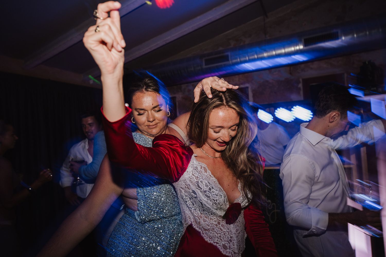 Wedding guests in colorful dresses dance energetically together under blue and red lights at lively wedding reception.