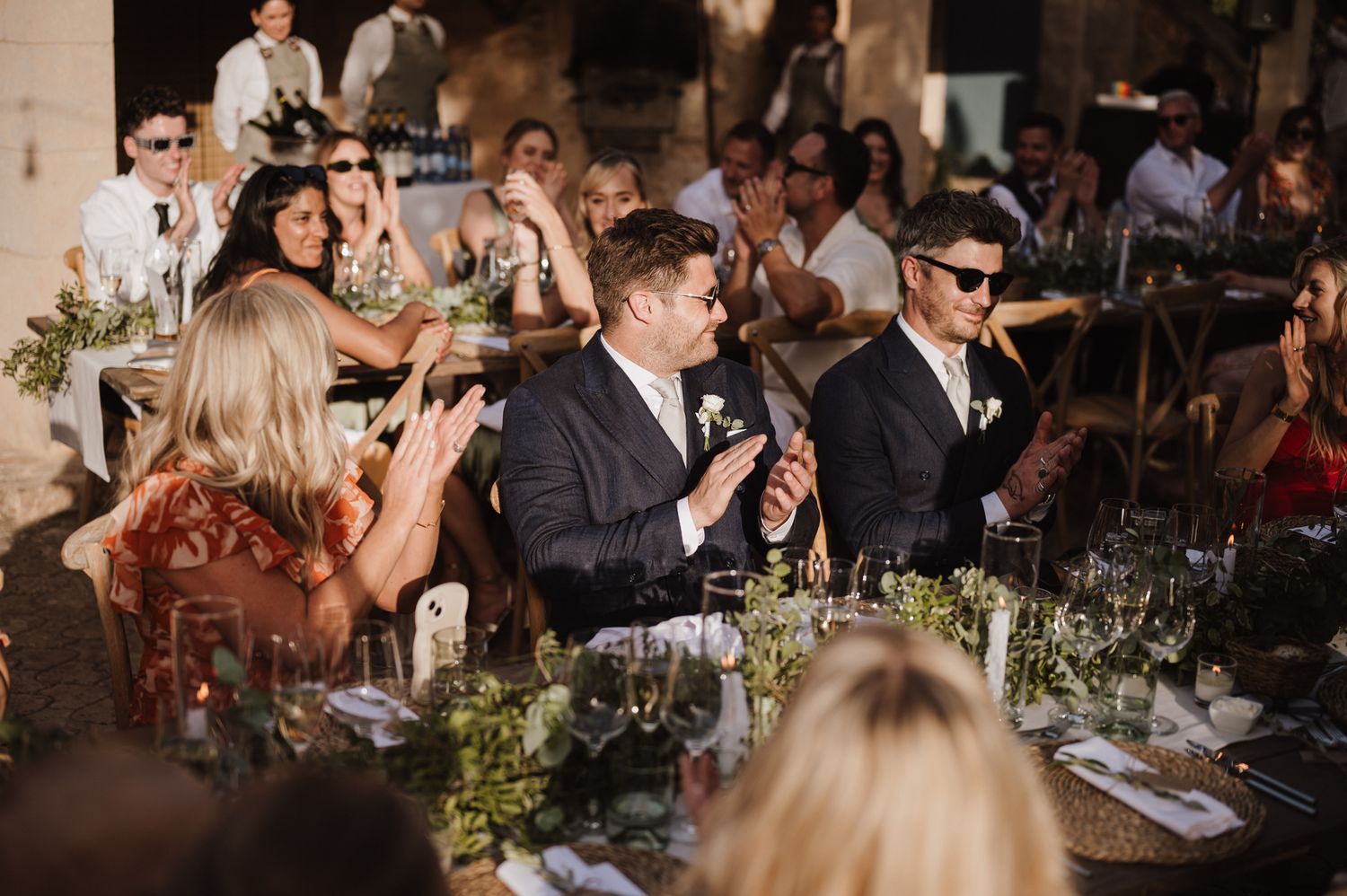 Wedding guests clapping and celebrating at a long decorated reception table in a rustic outdoor venue.