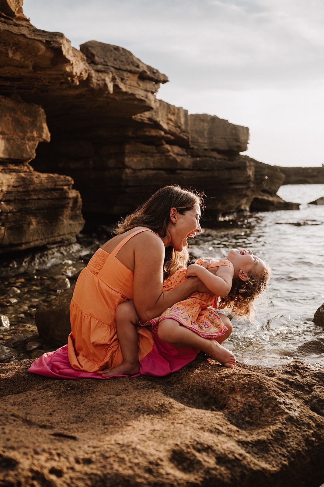 A mother laughing with her young daughter while sitting on rocky coastal cliffs by the ocean.