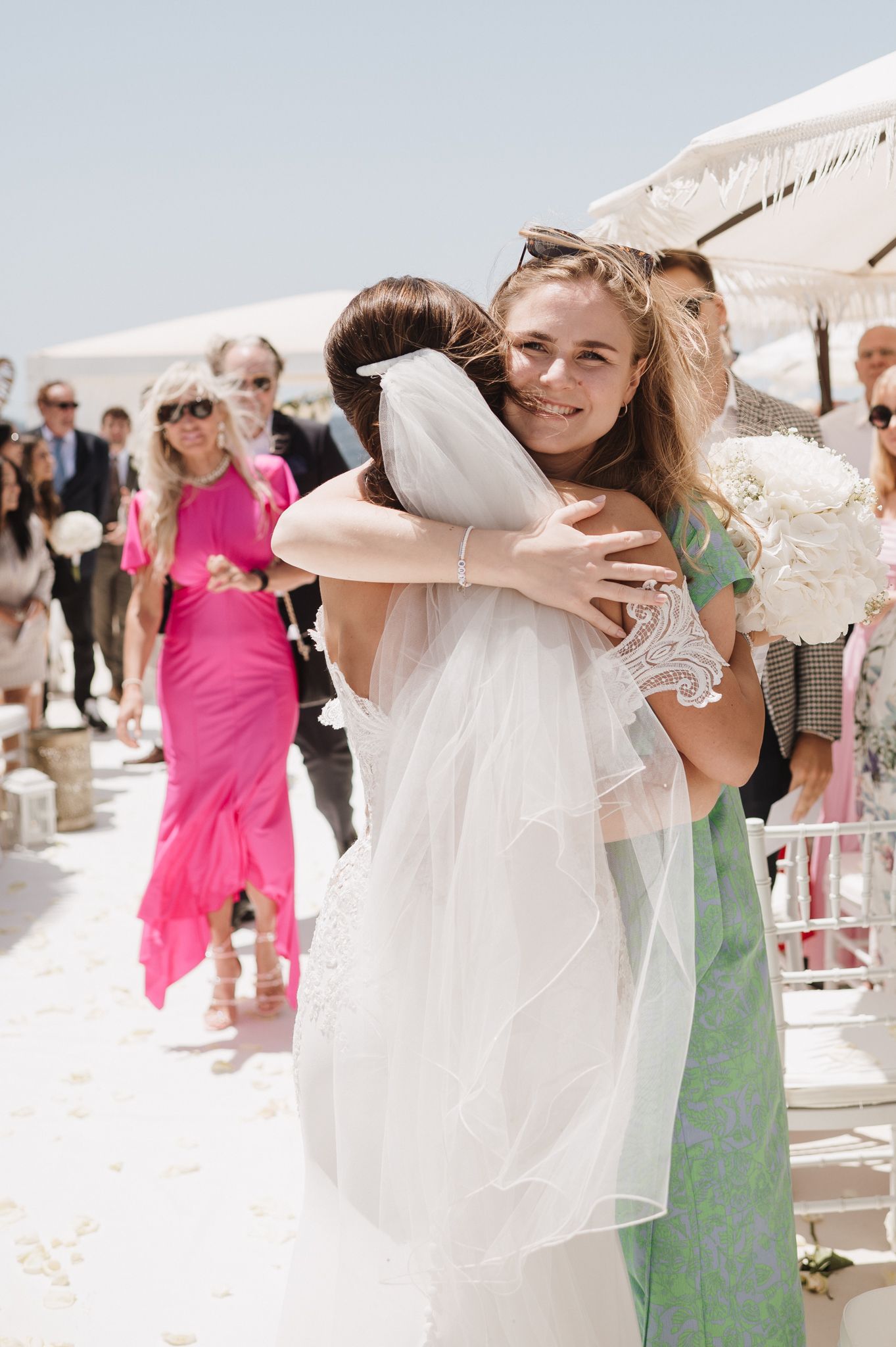 Bride in white lace gown and veil receives a warm hug from a guest during the ceremony.