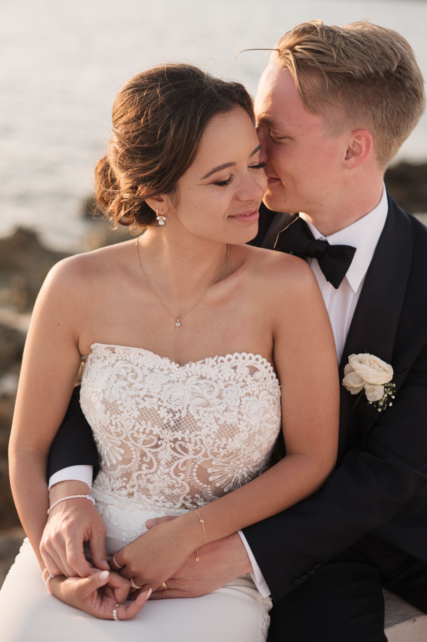 Romantic bride and groom portrait at sunset by the sea, groom in tuxedo nuzzling brides cheek.