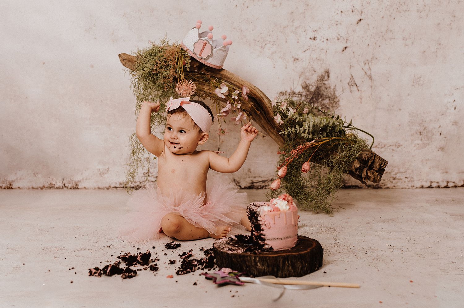 Baby girl in pink tutu sits beside smashed pink cake with floral decor during first birthday cake smash photo session.