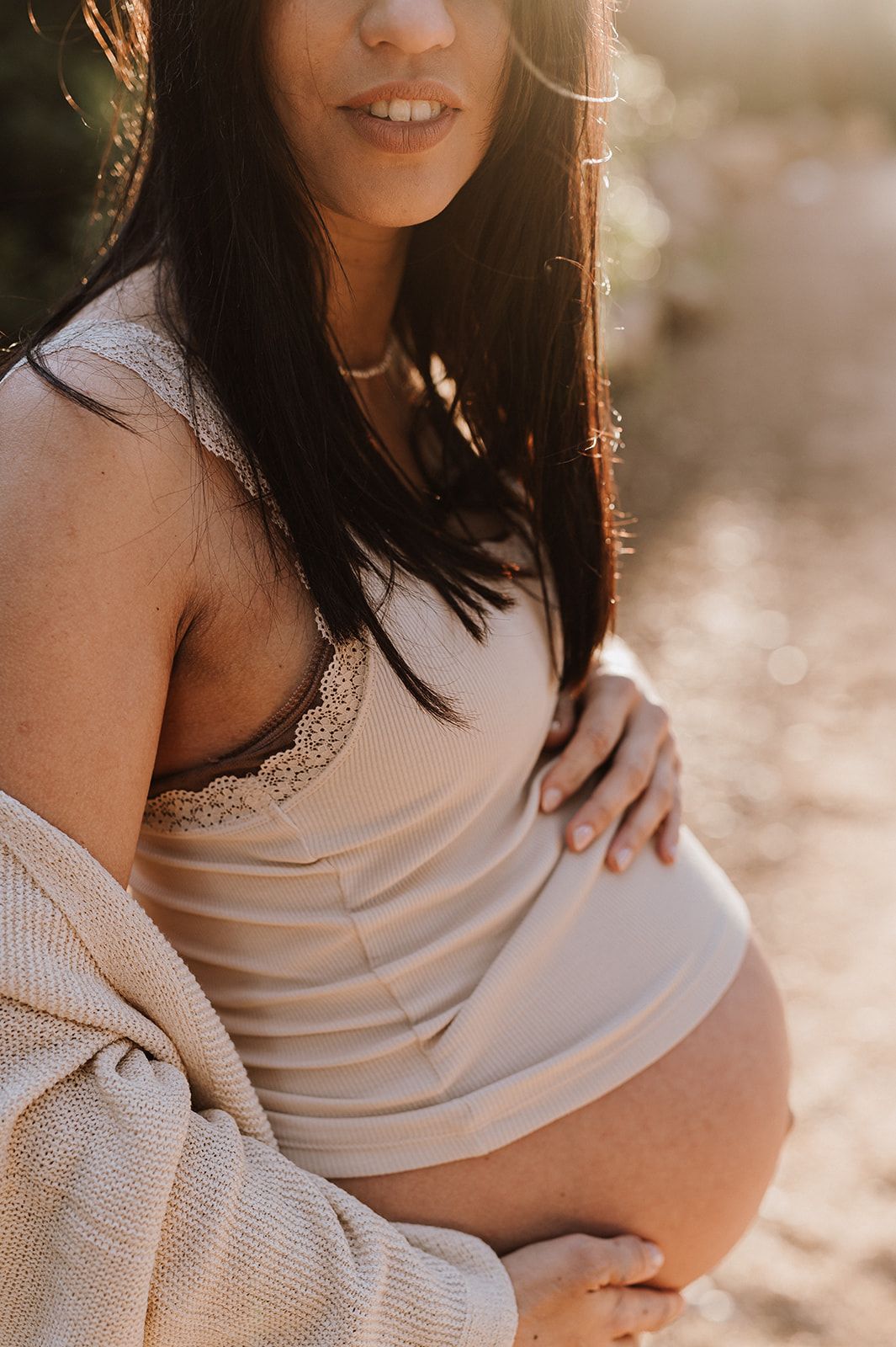 Pregnant woman in a beige striped dress and lace bralette cradling her baby bump outdoors in warm sunlight.