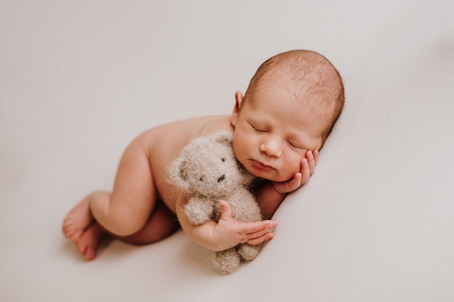 Peaceful newborn baby sleeping nude while cuddling a small teddy bear on a soft white background.