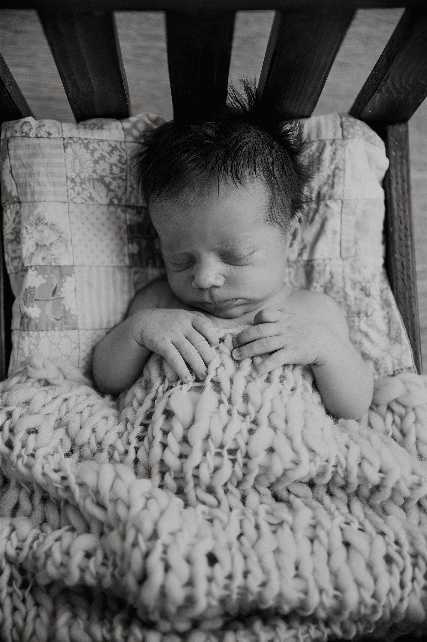 Black and white photo of a sleeping newborn baby wrapped in a chunky knit blanket inside a wooden crib.